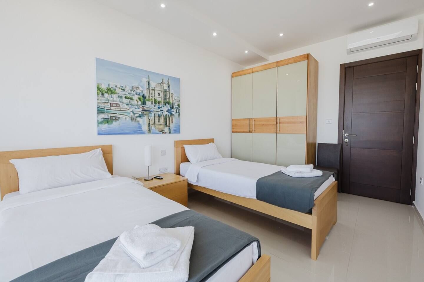 Habitación de hotel con dos camas, un espejo grande, un cuadro de un paisaje urbano y una puerta de madera oscura.