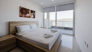 A modern bedroom with a large bed, a wooden headboard, a painting on the wall, and a balcony with a view of the sea. - This apartment is a 2 bedroom(s), 1 bathrooms, located in Il-Mellieħa, null. (Il-Mellieħa)