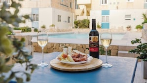 A table with a bottle of wine, two glasses, and a plate of food by a poolside with a view of buildings in the background.