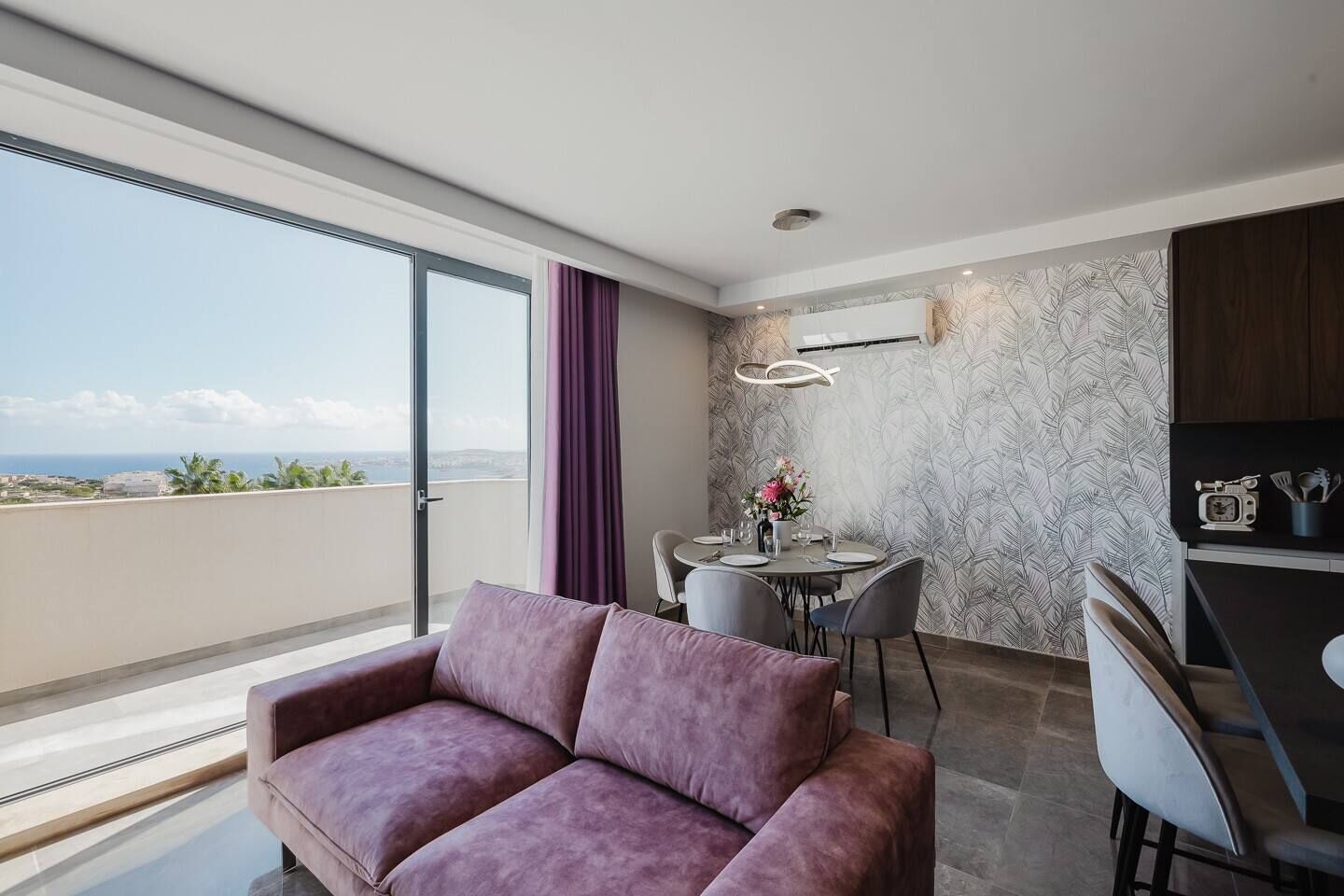 A modern room with a purple sofa, a dining table with chairs, and a kitchen area with a microwave and utensils.