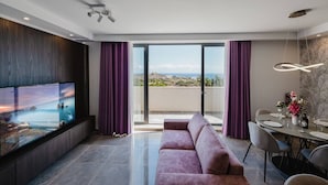 Un salón moderno con un televisor de pantalla plana grande, un sofá, una zona de comedor con una mesa preparada para dos personas, y un balcón con vistas panorámicas.