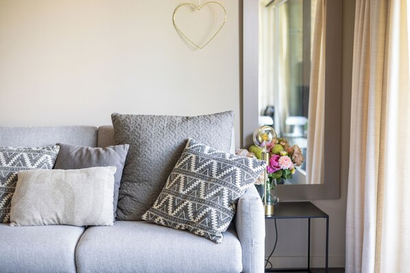 A grey sofa with patterned cushions, a small table with a vase of flowers, and a heart-shaped decoration on the wall.
