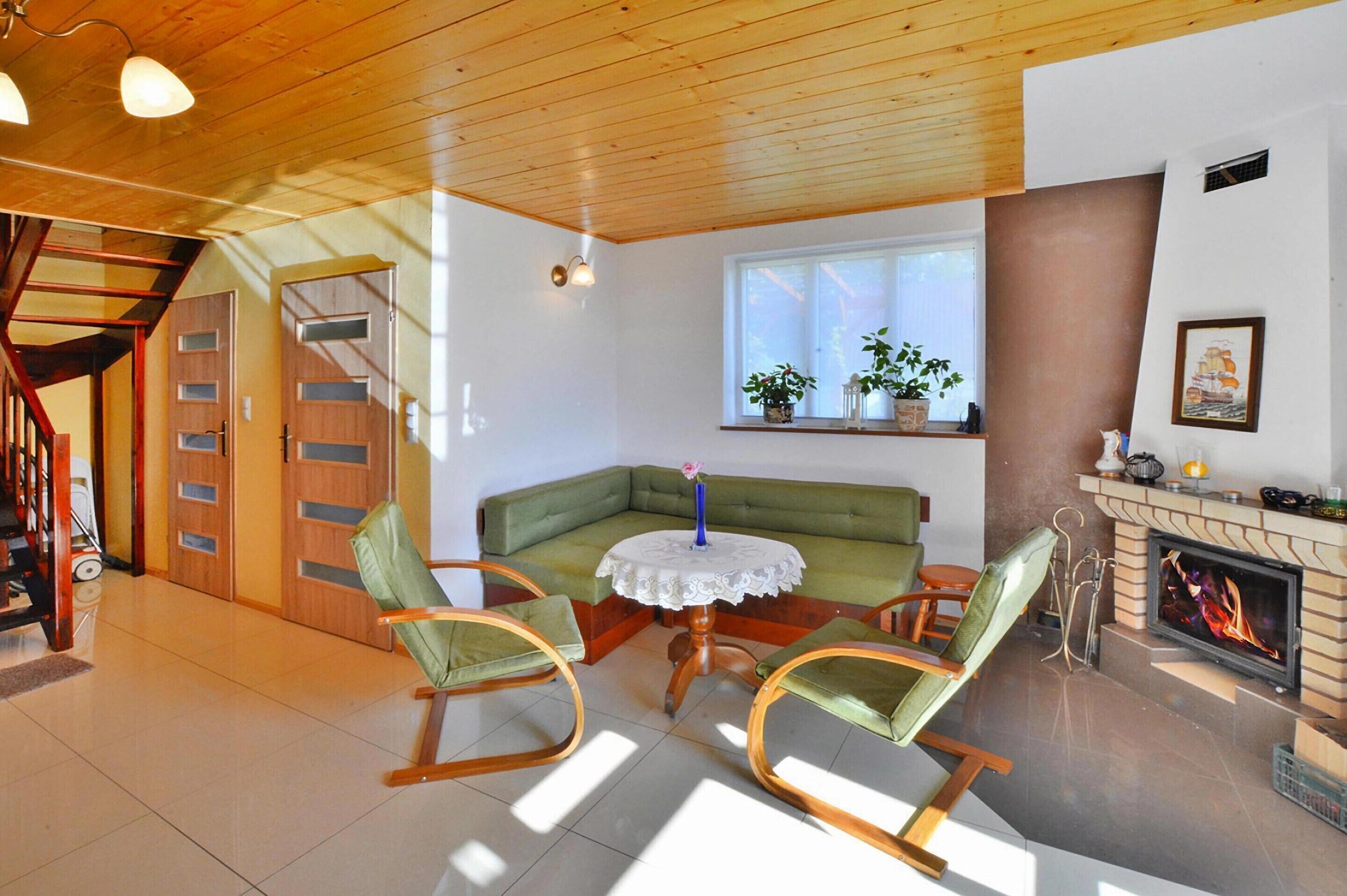 Un salon chaleureux avec un canapé vert, deux fauteuils à bascule, une petite table ronde, une cheminée et un escalier menant à une autre pièce.