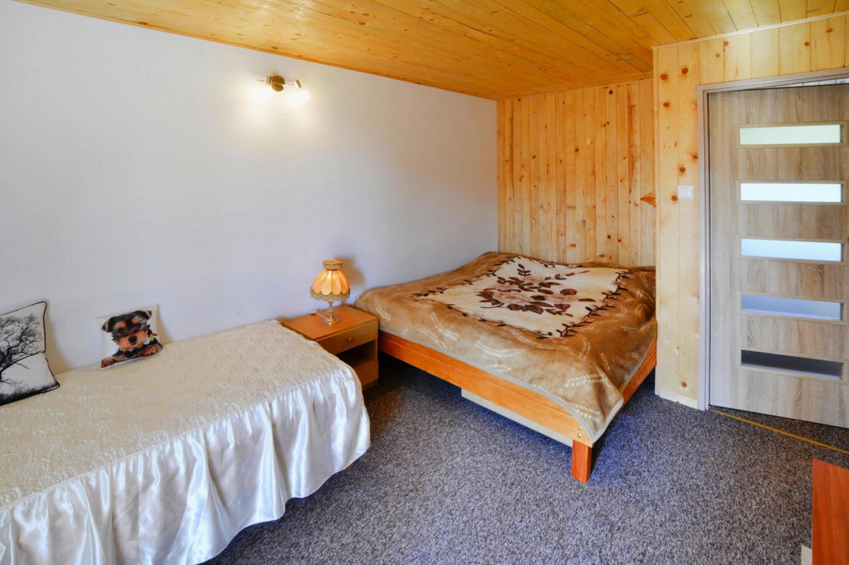 Un dormitorio con techo de madera, una cama con manta marrón, una mesita de noche con lámpara y un mueble auxiliar con un adorno de oso de peluche.