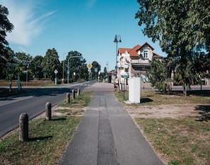 A paved sidewalk adjacent to a road with bollards, trees, and a building with a red roof. - Doppelhaushalfte, Schonwalde (Schönwalde-Glien)