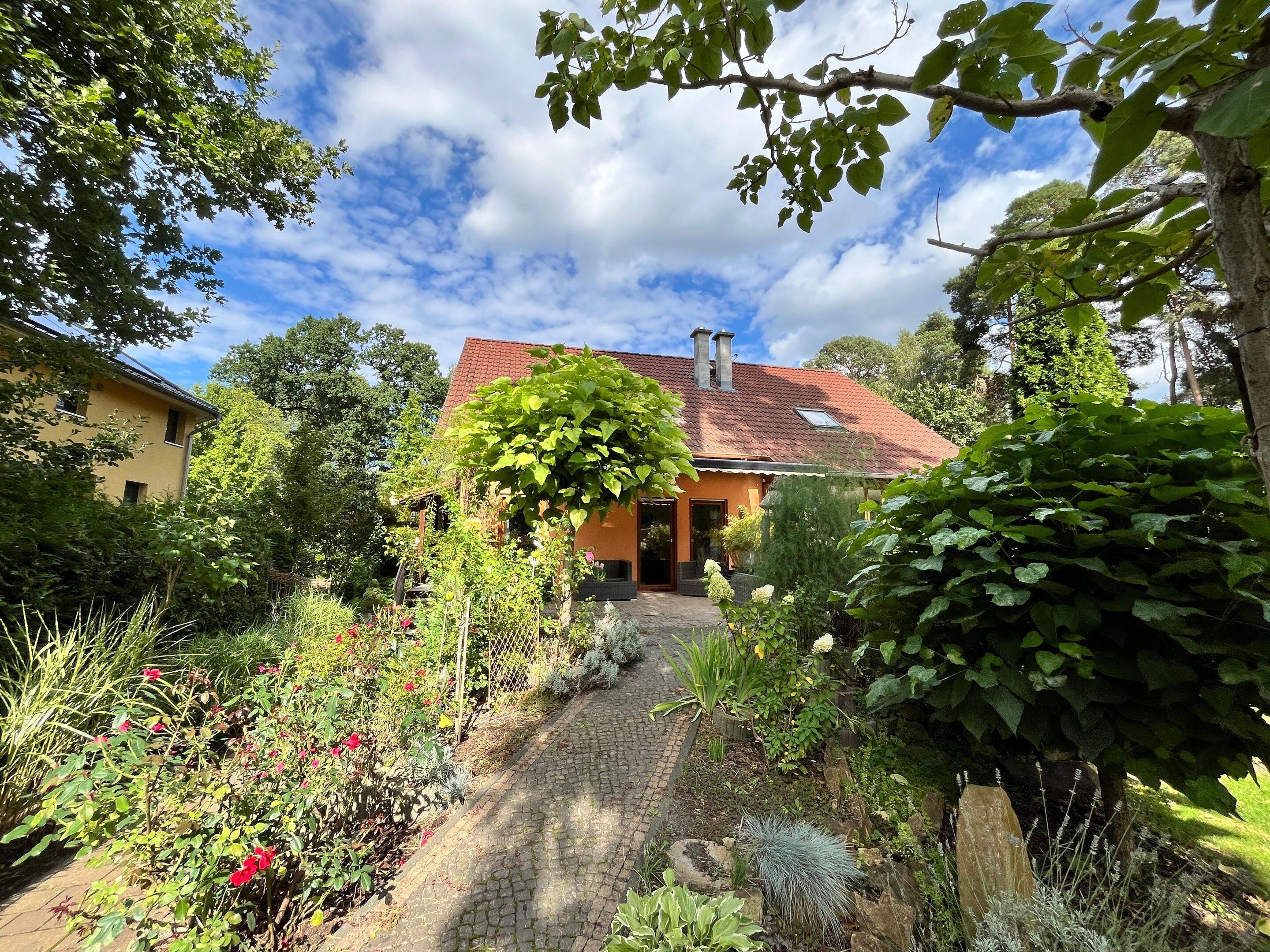 A house with a red-tiled roof, surrounded by a garden with various plants and a cobblestone path leading to the entrance.