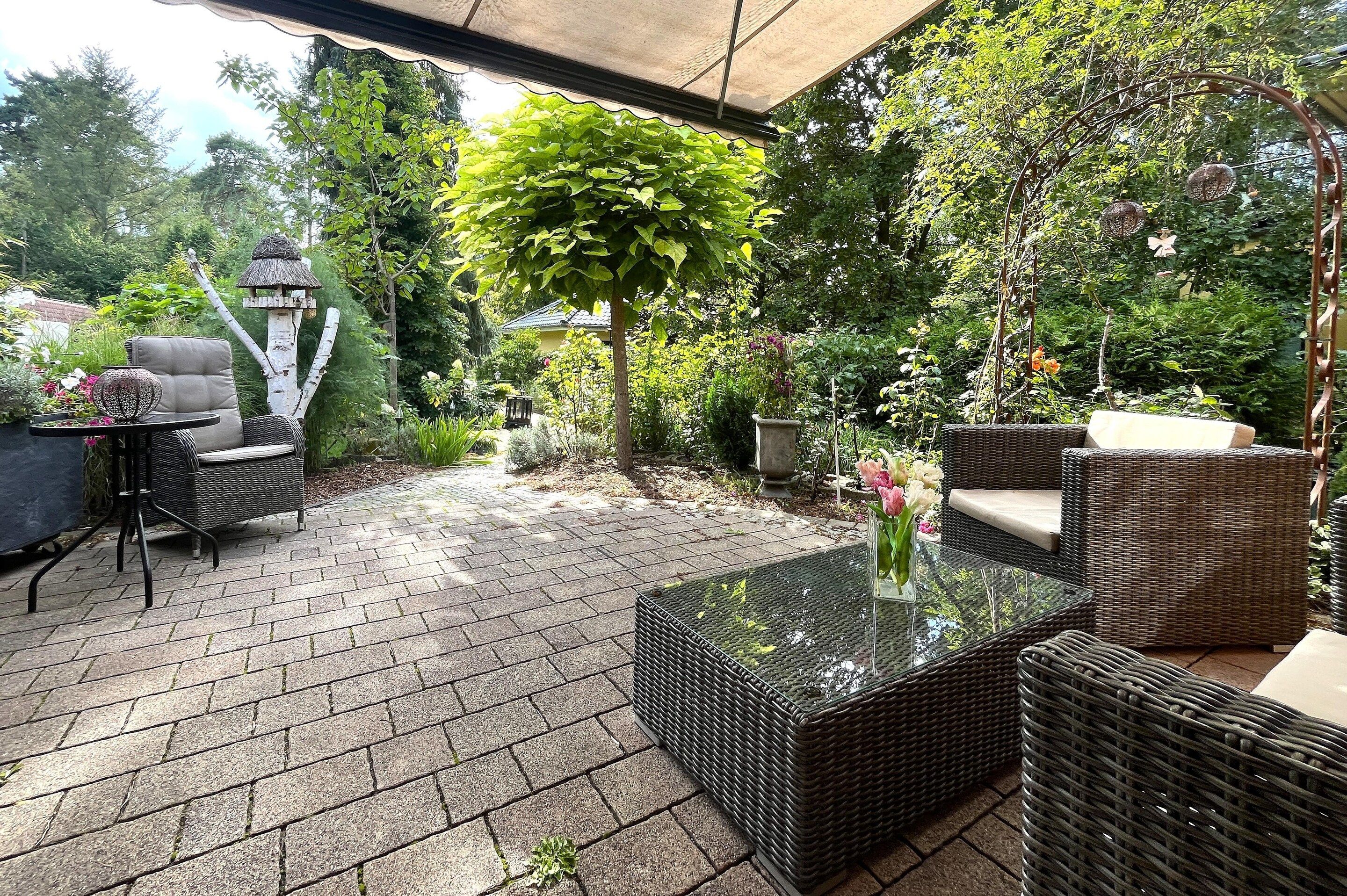 An outdoor patio with wicker furniture, a glass table, and a variety of plants.