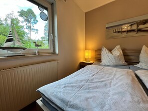 A hotel room with a bed, a nightstand with a lamp, a window with a view of trees, and a radiator. - Doppelhaushalfte, Schonwalde (Schönwalde-Glien)