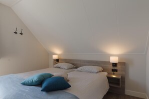 Een slaapkamer met een schuine plafond, twee nachtkastjes met lampen, een bed met een lichtgékleurd dekbed en kussens, en een houten hoofdbord.