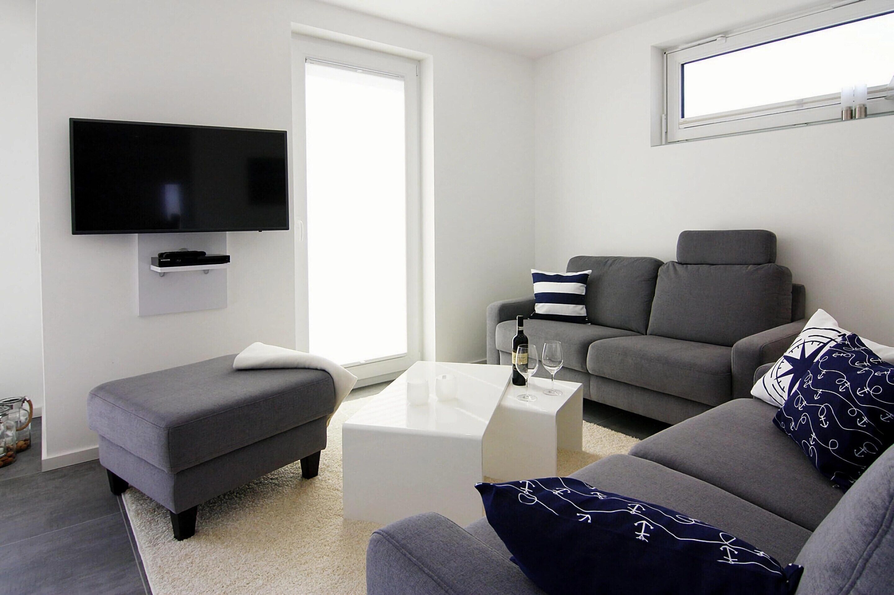 Een moderne woonkamer met een flatscreen televisie aan de muur, een grijze bank, een witte salontafel en een grijze fauteuil.