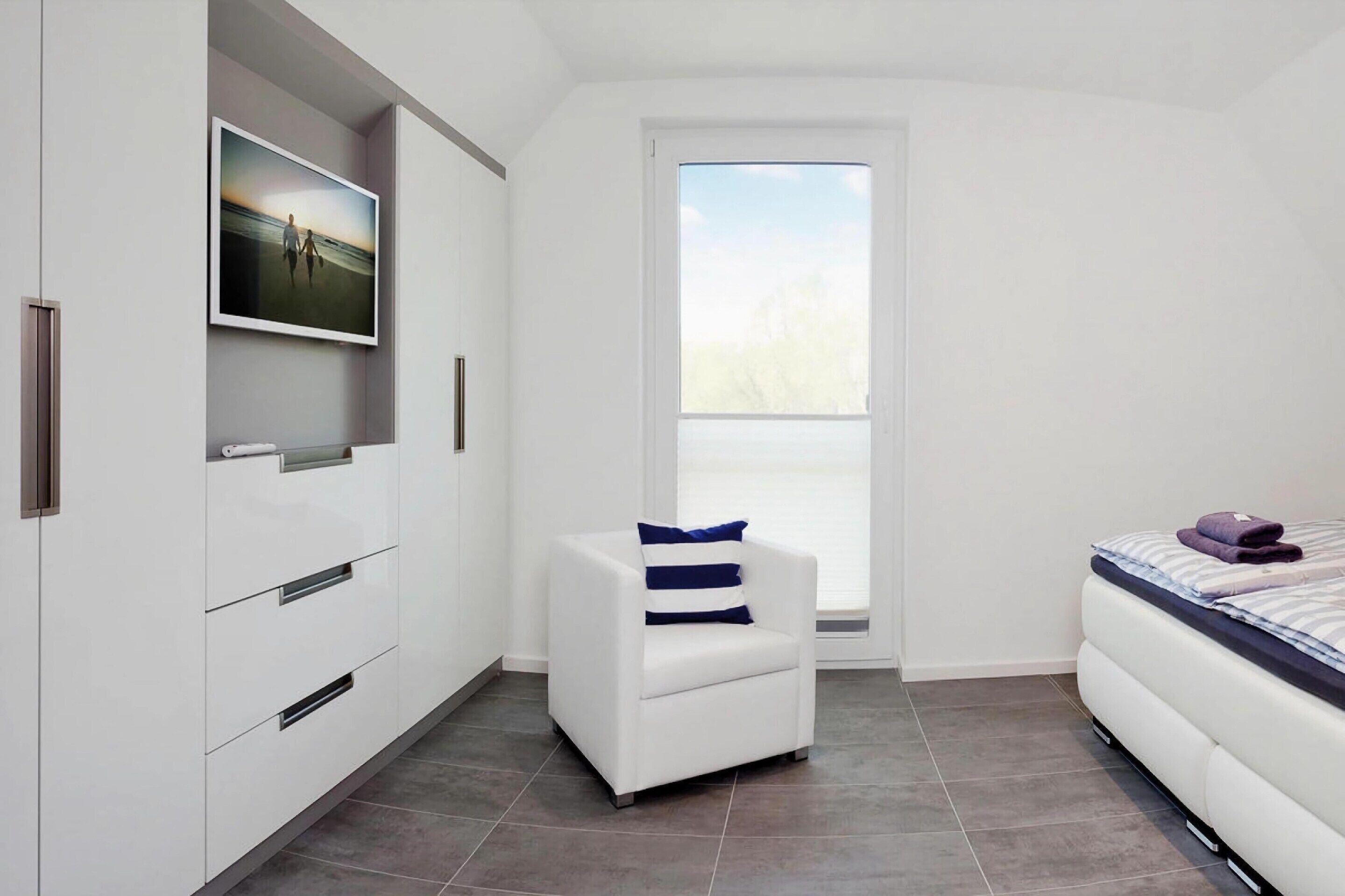 A modern bedroom with a white wardrobe, a white armchair with a striped pillow, a bed with a striped duvet, and a large window with a view of trees.
