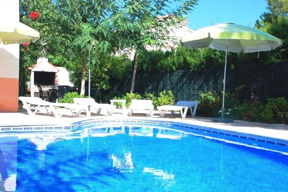 Une piscine entourée de verdure, avec des chaises longues blanches et des parasols, et un bâtiment visible à l’arrière-plan.