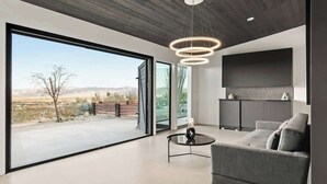 Un salon moderne doté d’une grande porte-fenêtre coulissante donnant sur une terrasse extérieure, d’une table basse ronde, d’un canapé gris et d’un luminaire suspendu contemporain.