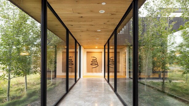 Un couloir aux murs de verre, au plafond en bois et au sol en béton, qui mène à une porte ornée d’un logo.