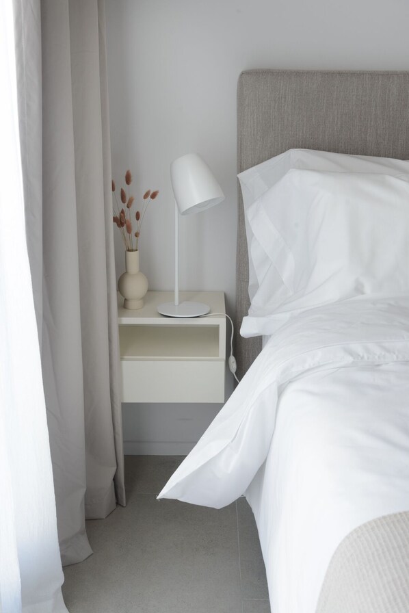 A minimalist hotel room with a bed, a bedside table with a lamp, and a nightstand with a vase of dried flowers.