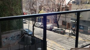 Varanda com cadeiras de vime e uma pequena mesa redonda, com vista para uma rua com carros estacionados e edifícios.