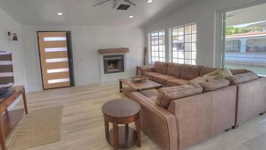 Ein Wohnzimmer mit Ecksofa, Kamin, Deckenventilator und großen Fenstern.