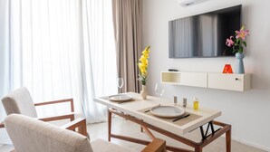 Un salon moderne avec une table à manger pour deux personnes, un canapé, un téléviseur fixé au mur et une grande fenêtre avec des rideaux.