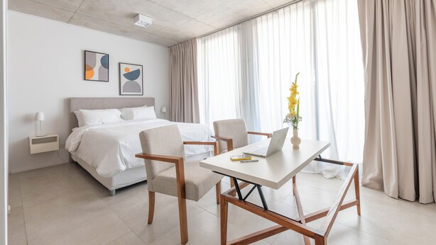 Une chambre d’hôtel moderne, équipée d’un lit, de deux fauteuils, d’une table avec un ordinateur portable, d’un vase de fleurs et d’œuvres d’art accrochées au mur.