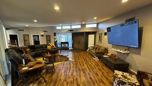 A spacious living room with a large flat-screen TV mounted on the wall, a leather sofa, a wooden coffee table, and a rustic wooden floor. - Dock Holiday – The Keys Favorite | Wild West River Escape (Parker)