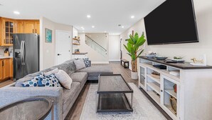 Ein modernes Wohnzimmer mit Sofa, Couchtisch und Fernseher. Eine Küchenzeile mit Holzausstattung und Kühlschrank ist ebenfalls vorhanden. Im Hintergrund ist eine Treppe zu sehen.