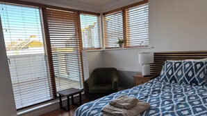 Una habitación de hotel con una cama, una silla, una mesita de noche con lámpara y ventanas amplias con persianas.