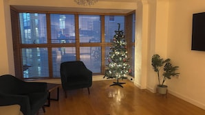 Una sala de estar con piso de madera, un sillón negro, una mesita, un árbol de Navidad y un ventanal grande con persianas.