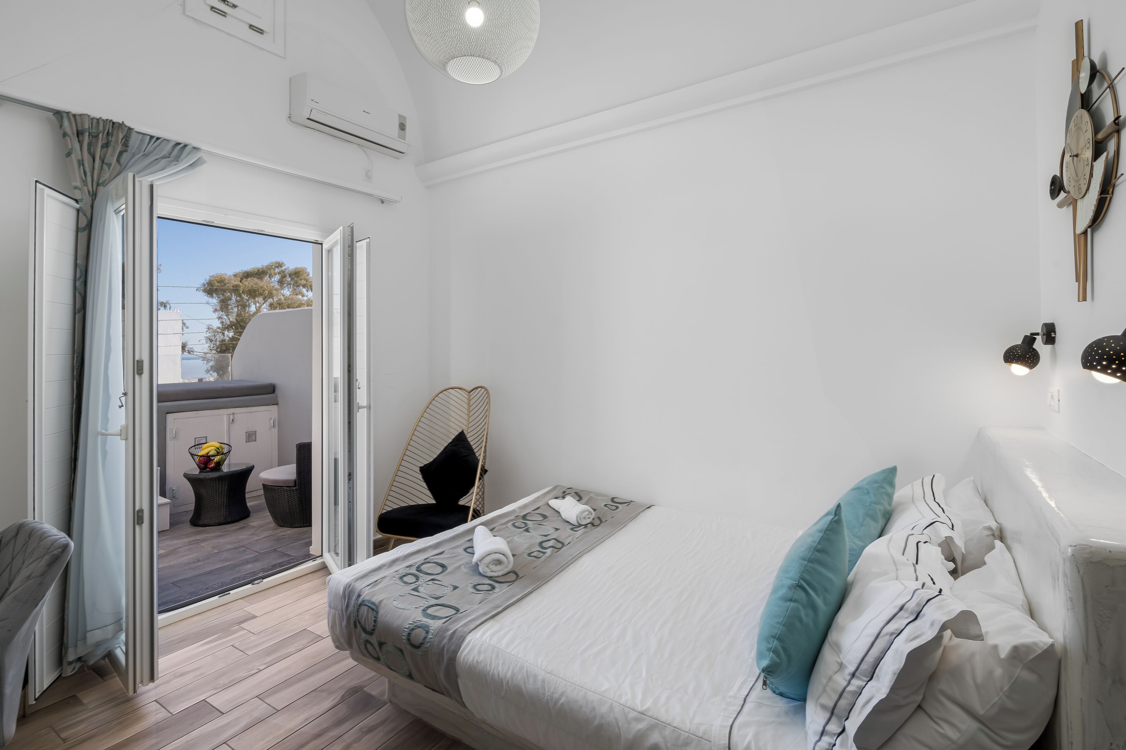 A bedroom with a bed, a chair, a small table with fruit, and a view through an open door to an outdoor area.