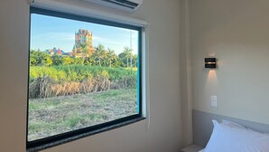 Una habitación de hotel con un ventanal grande, un aire acondicionado, una mesita de noche y una lámpara fijada a la pared.