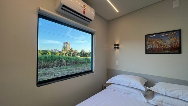 Una habitación de hotel con un ventanal grande, un equipo de aire acondicionado, una cama con sábanas blancas, una lámpara fijada a la pared y un cuadro enmarcado.