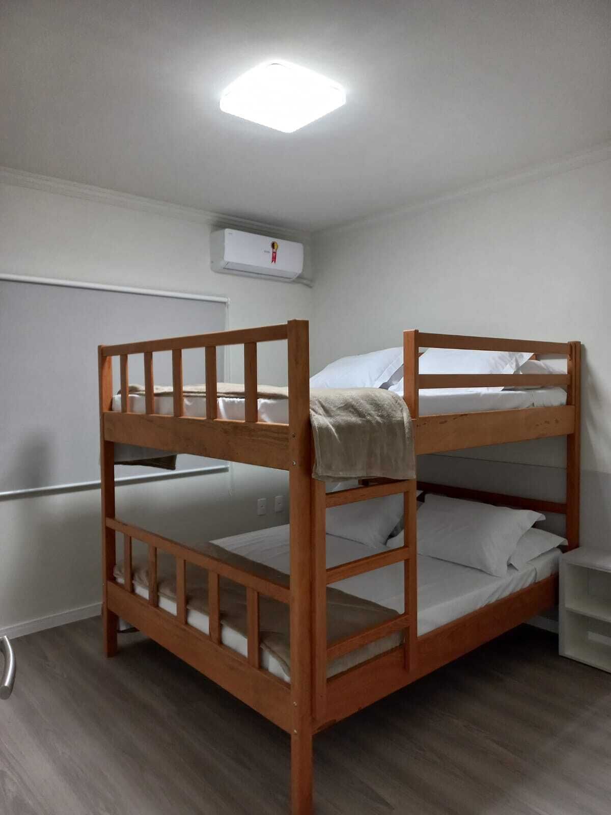 A bunk bed with a wooden frame and white bedding, a white wall, and a ceiling light.