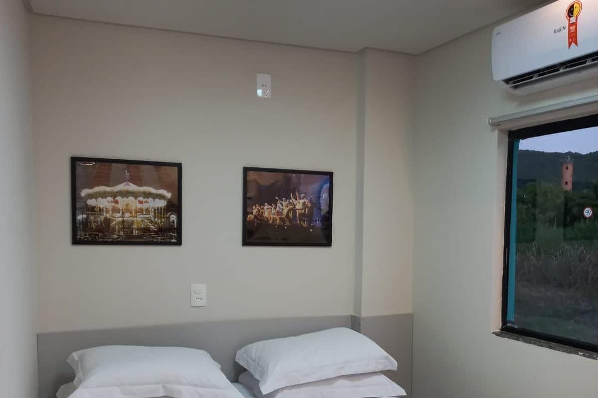 A hotel room with a bed, two framed pictures on the wall, a window with a view, and an air conditioning unit.