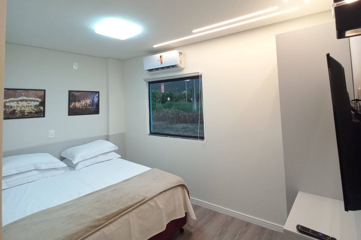 A modern hotel room with a bed, a flat-screen TV, and a window with a view of greenery.