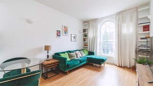 Ein Wohnzimmer mit einem grünen Sofa, einem Glastisch, einem Beistelltisch und einem Bücherregal.