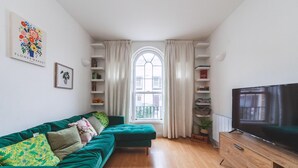 Ein Wohnzimmer mit einem grünen Sofa, einem Fernseher, einem Bücherregal und einem Fenster mit Vorhängen.
