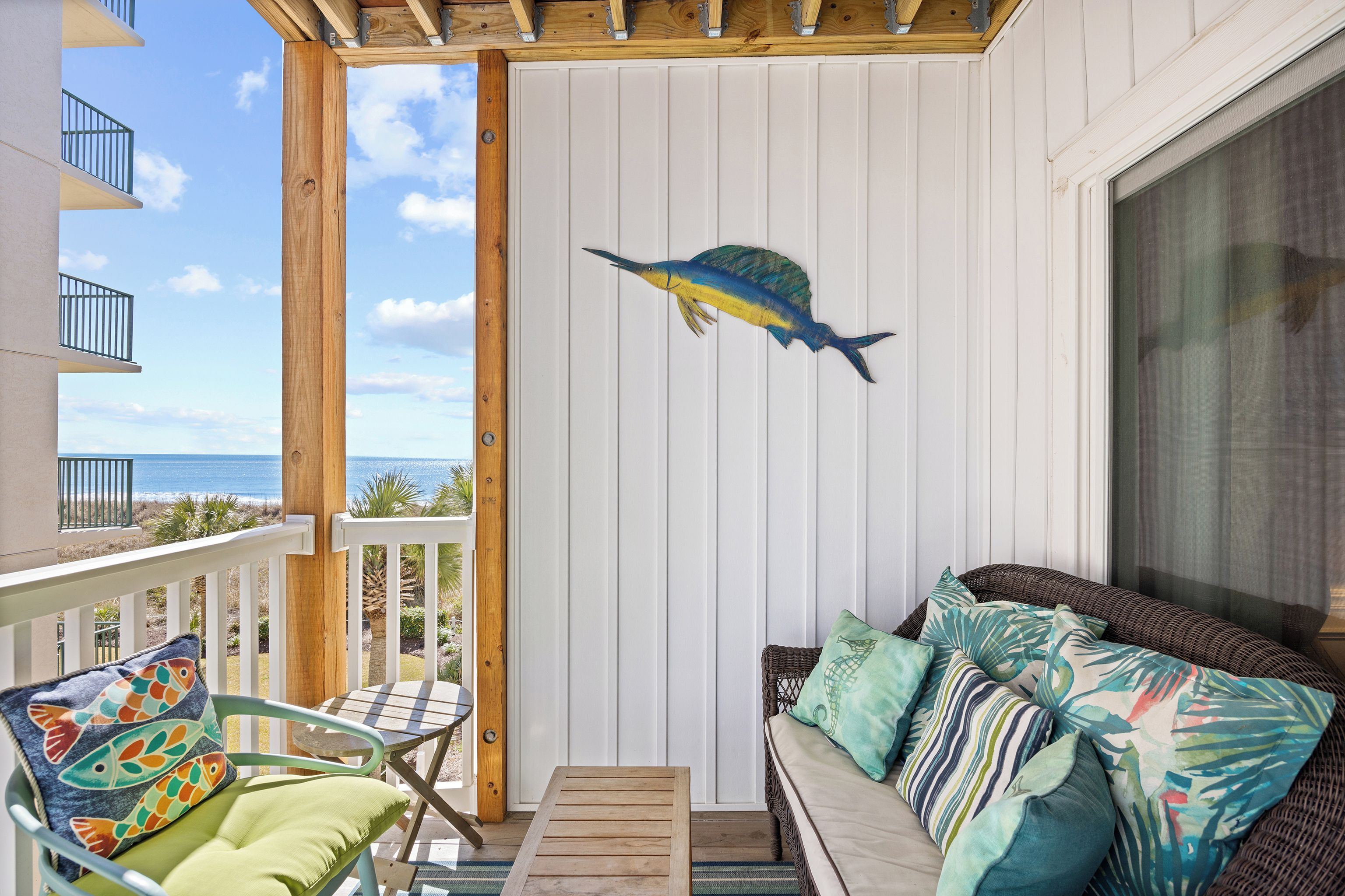 A balcony with a view of the ocean, a wicker sofa with patterned cushions, and a wooden table.