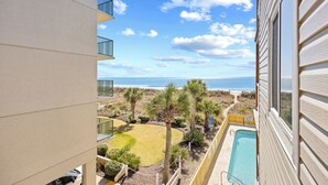 Un balcone con vista sulla spiaggia e sull'oceano, una piscina e un cortile recintato con palme.