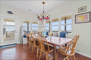 A dining area with a wooden table and chairs, a chandelier, and large windows.