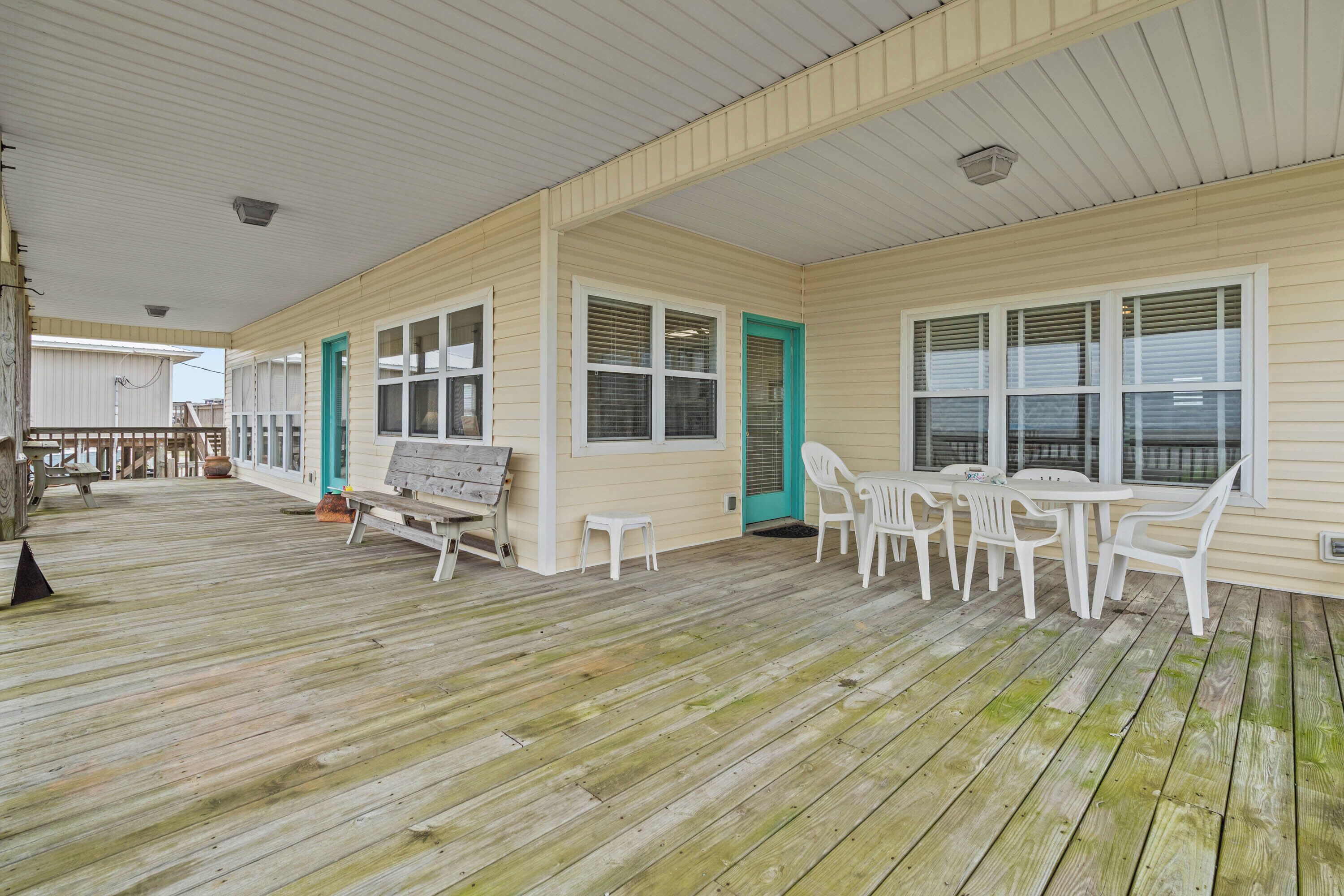 A covered deck with wooden flooring, white plastic chairs, and tables, and a door with a green frame.