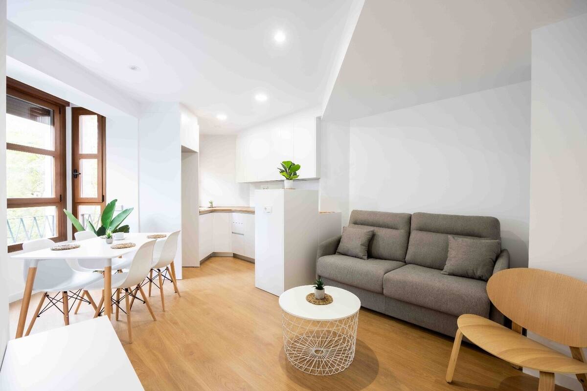 Una sala de estar moderna con un sofá gris, una mesa redonda y un comedor con una mesa y sillas blancas.
