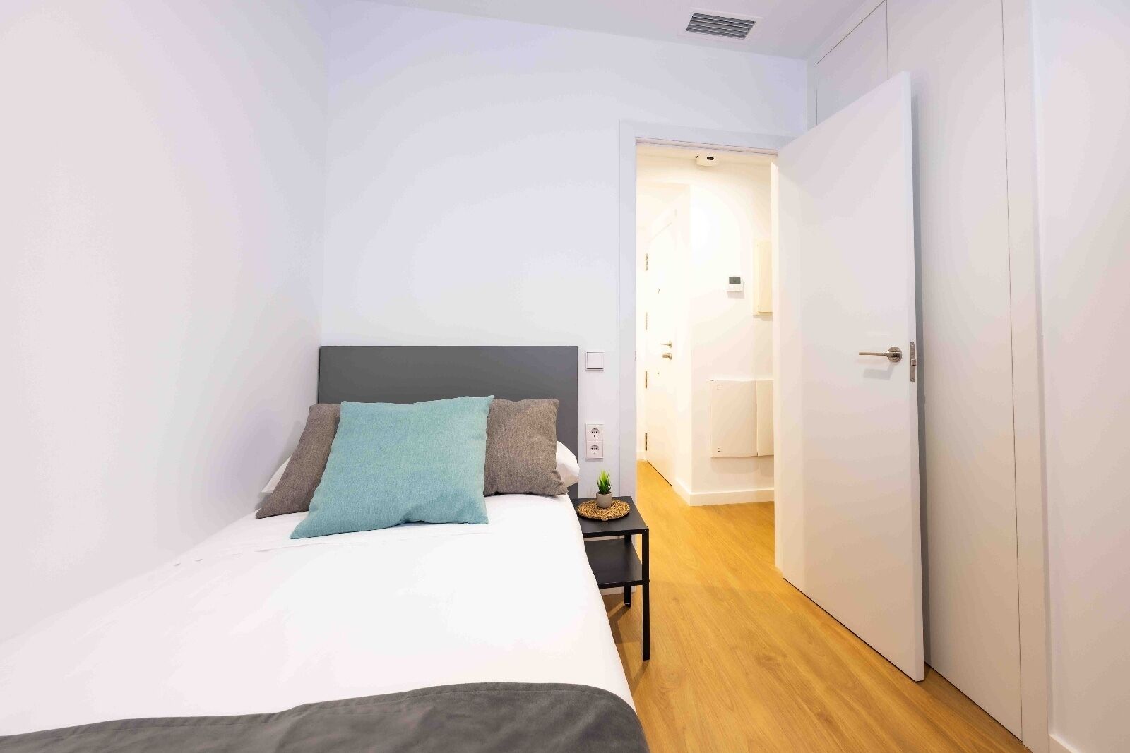 Un dormitorio con cama, una mesita de noche con lámpara, y una puerta que da a otra habitación.