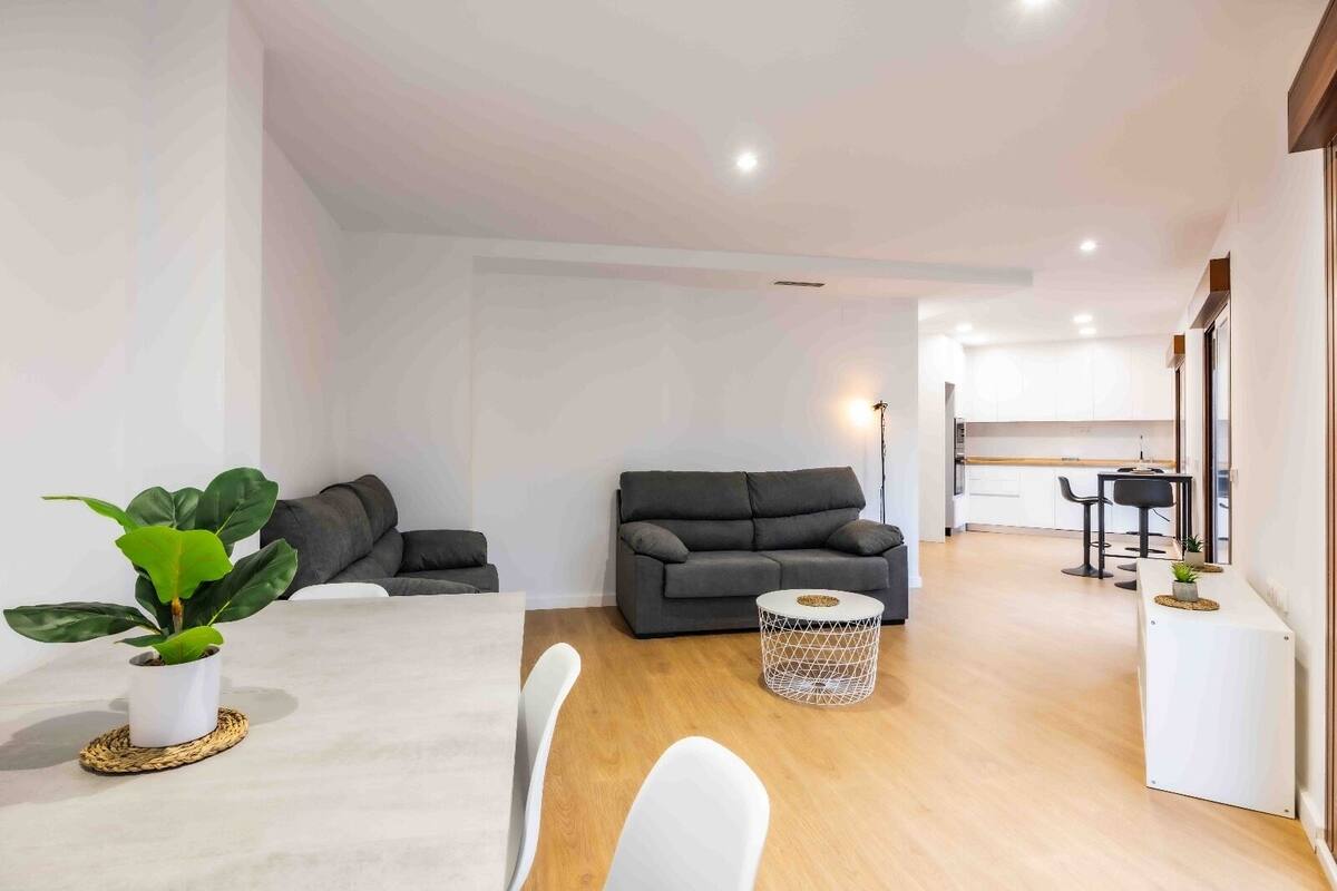 Una sala moderna con un sofá gris, una mesa de centro redonda y un comedor con una mesa y sillas blancas.