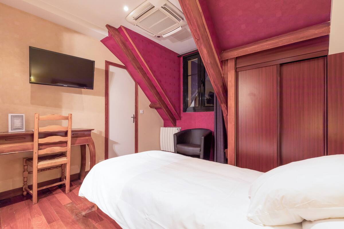 A bedroom with a bed, a desk with a chair, a television, and a window.