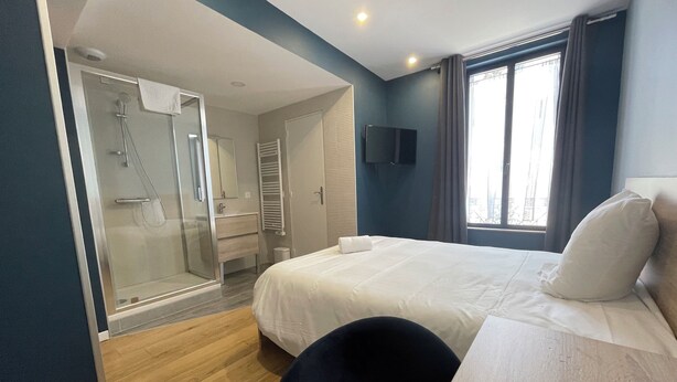 A hotel room with a bed, a desk, a chair, a TV, a shower area, and a window with curtains. - Hôtel Le Celtic (Le Thillay)
