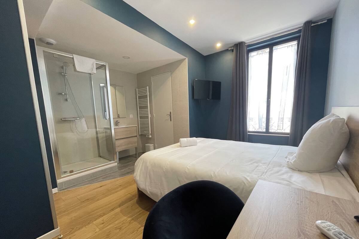 A hotel room with a bed, a desk, a chair, a TV, a shower area, and a window with curtains.