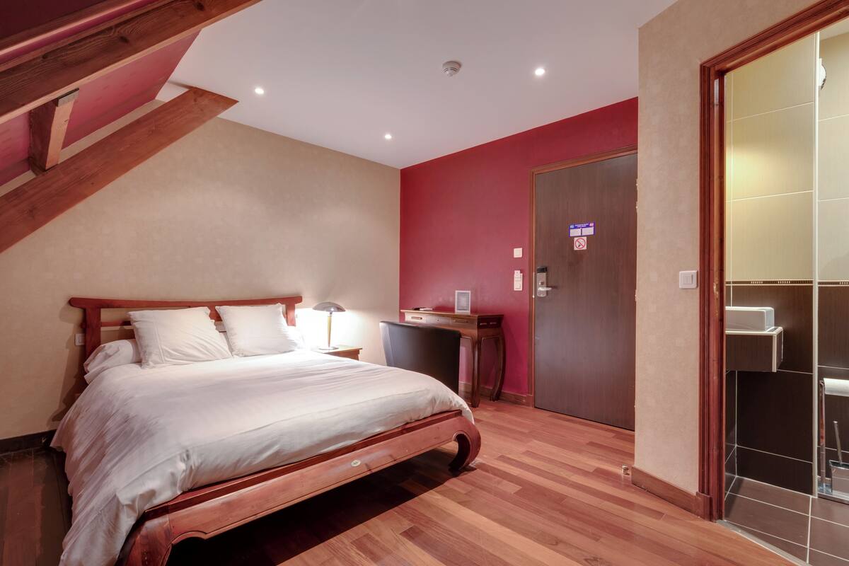 A bedroom with a wooden bed, a desk, and a bathroom with a glass door.