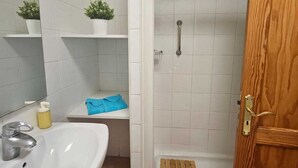 A bathroom with a white sink, a mirror, two potted plants on shelves, a shower with a wooden door, and a towel on the floor.