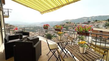 A balcony with wicker furniture, potted plants, and a view of a town and mountains.