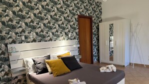 Una cama bien hecha con almohadas, una mesita de noche con botellas y un armario en una habitación luminosa con papel tapiz estampado.
