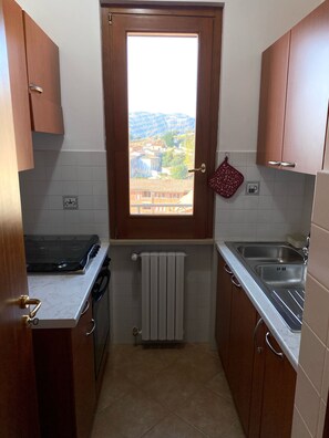 Une cuisine compacte avec des meubles en bois, une cuisinière à gaz, un four encastré, une fenêtre donnant sur un paysage vallonné et un torchon rouge accroché au mur.